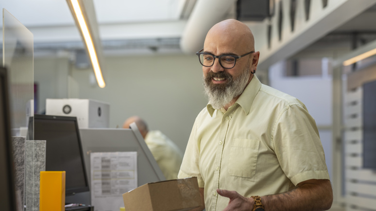 Ein Postbeamter mit Glatze, Brille und gelbem Hemd arbeitet am Schalter und hat ein Paket in der Hand